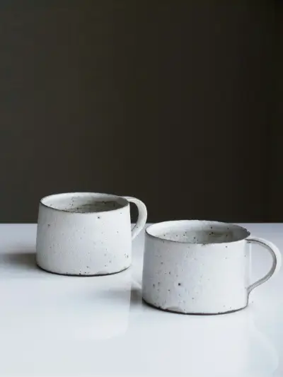 Two speckled white stoneware mugs with loop handles, on a white table against a dark wall.